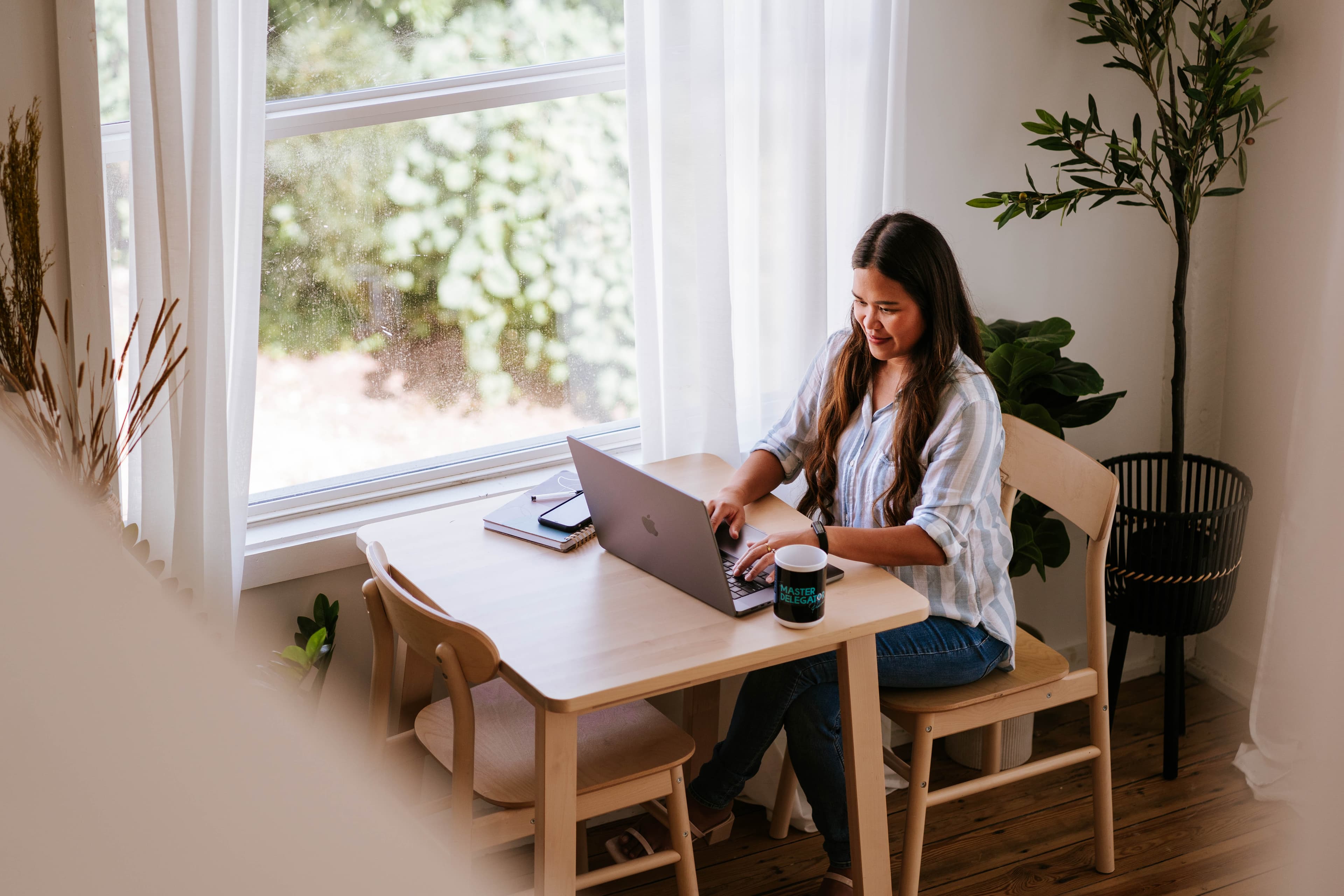 Kristy Yoder working on a laptop at a sunlit table with a “Master Delegator” mug beside her