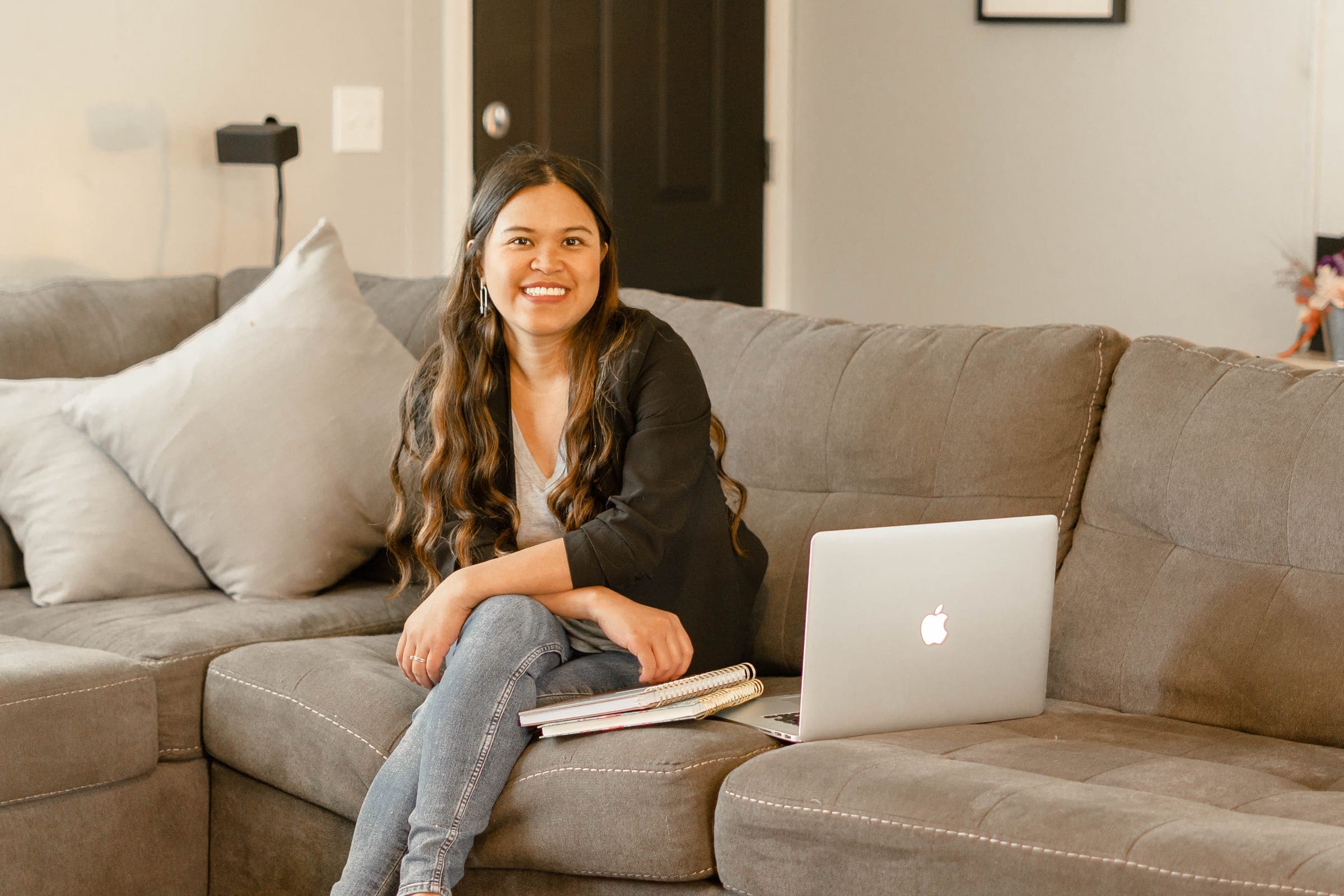 Kristy Yoder smiling while seated on a gray couch with a laptop and notebooks beside her