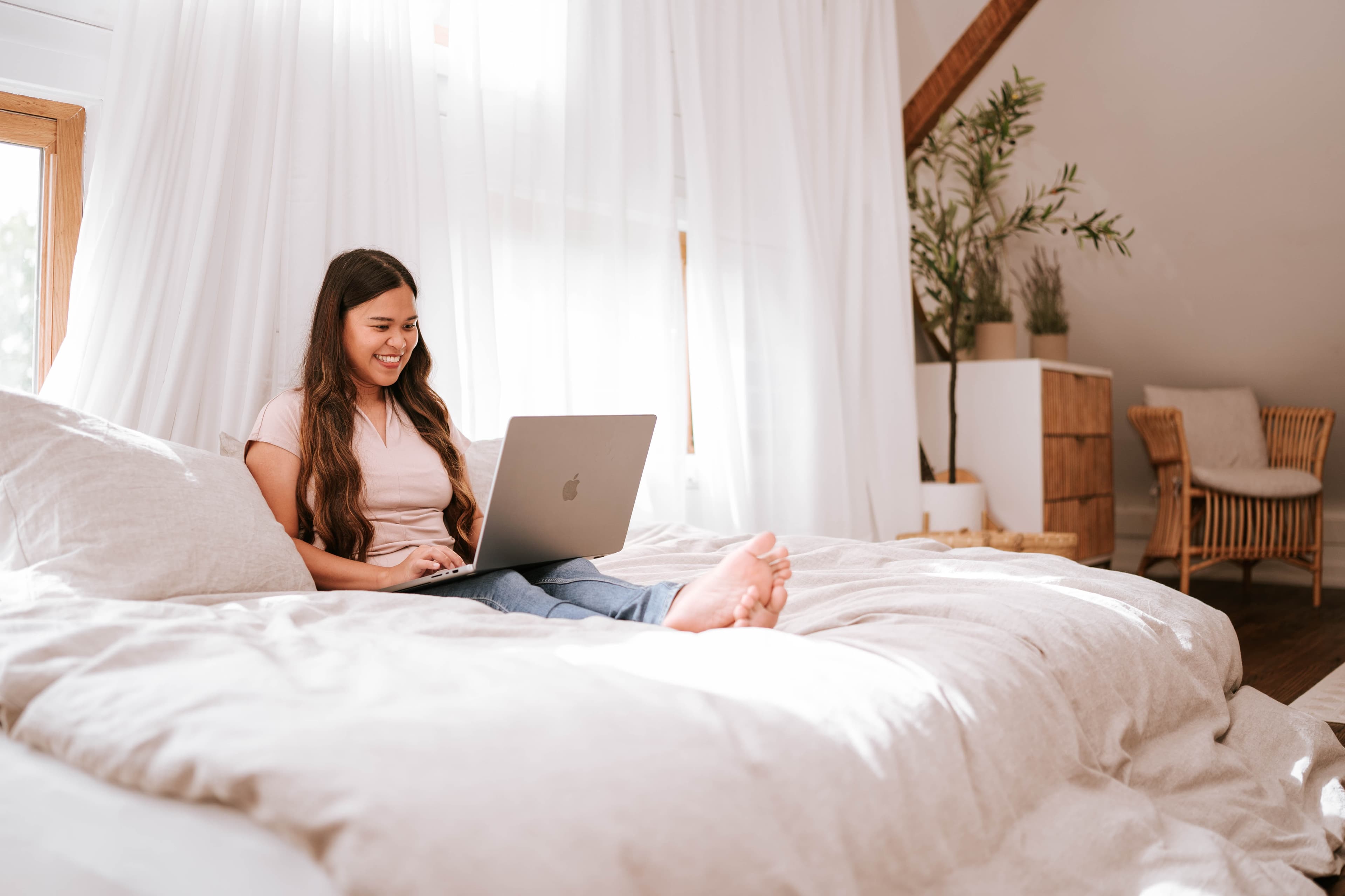 A woman smiling while working on her laptop in a bright, calm bedroom