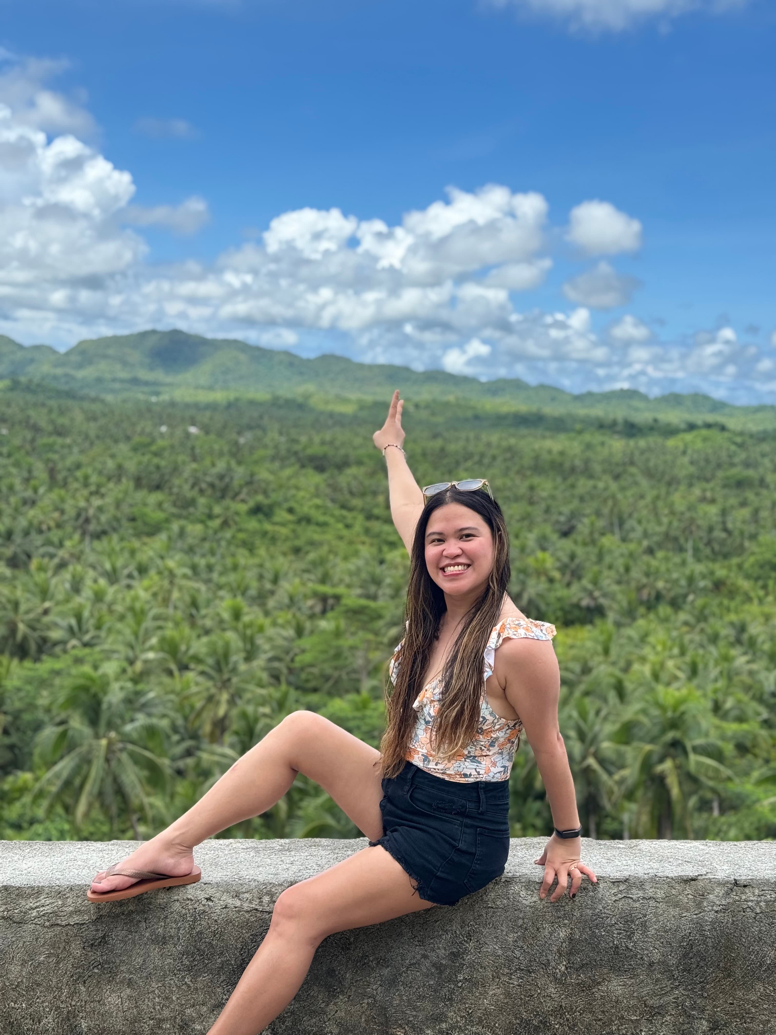 Kristy Yoder smiling with one arm raised while sitting on a ledge overlooking a green palm-tree valley