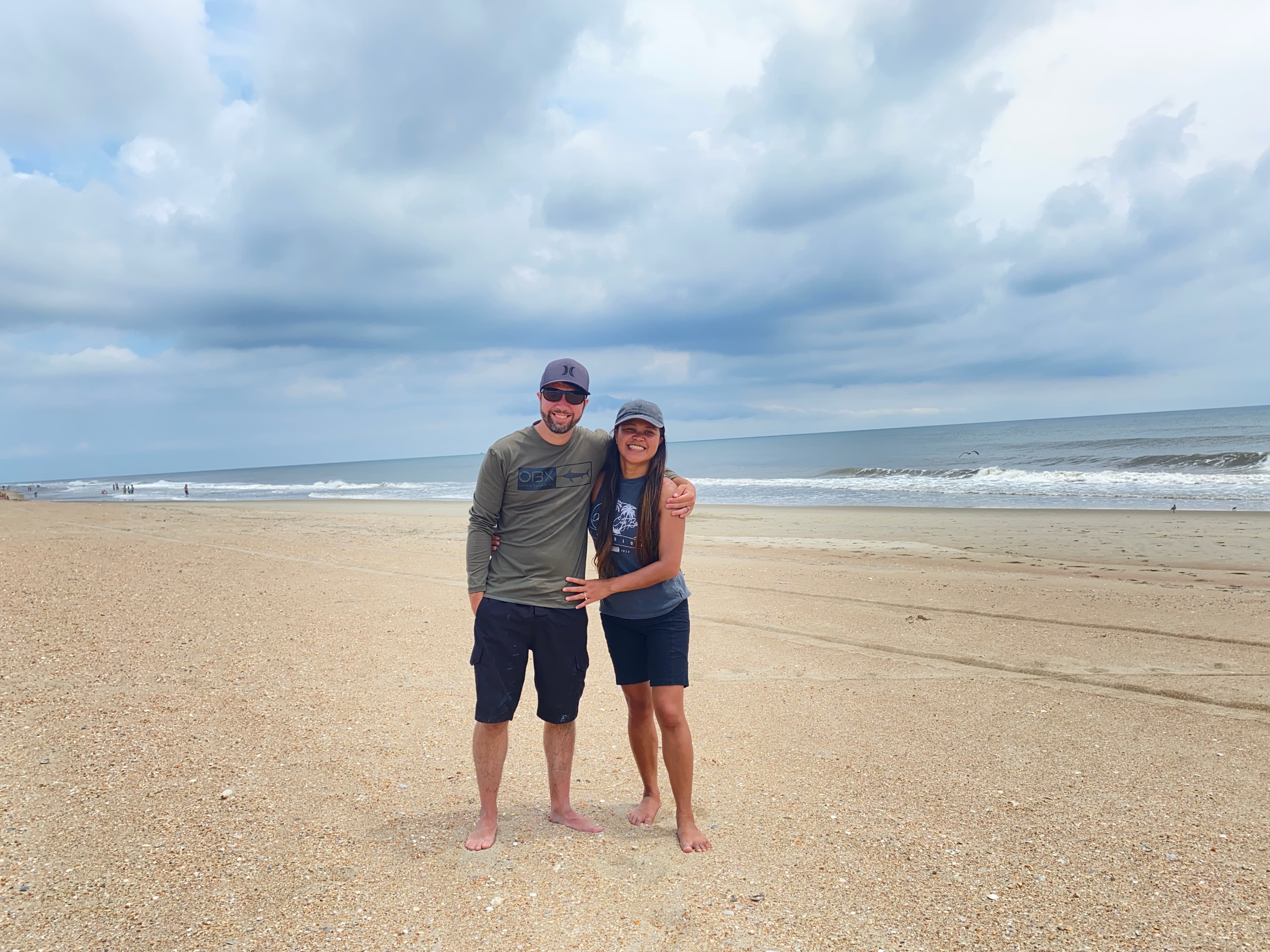 Kristy Yoder and her husband standing together barefoot on a wide beach under a cloudy sky